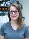 A white woman with light brown shoulder-length hair and black-rimmed glasses wears a gray v-neck t-shirt and smiles at the camera in front of a laboratory shelf.